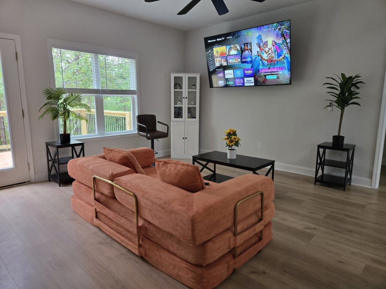 A living room with a sectional sofa, a TV mounted on the wall, and a window with blinds.
