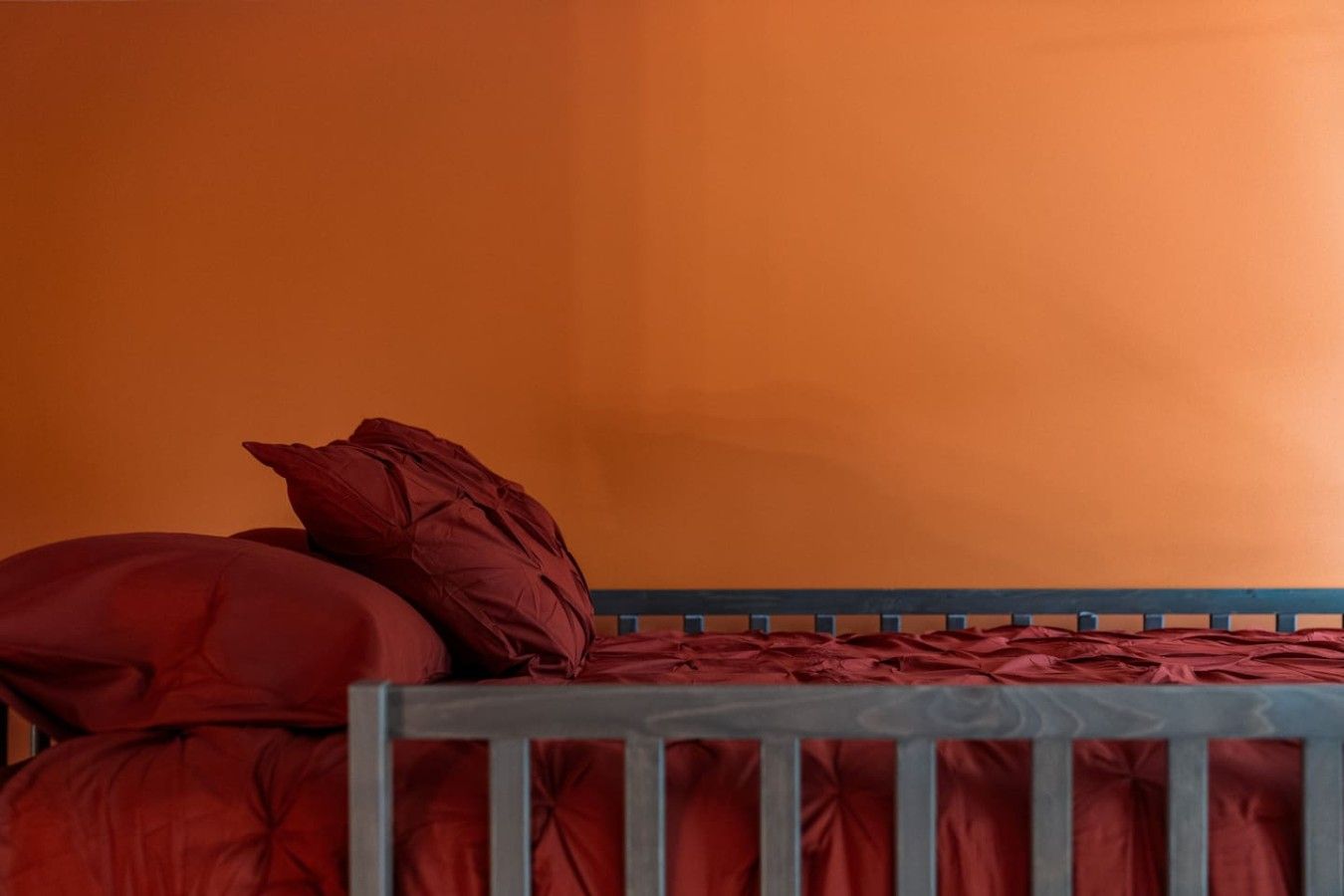 A single bed with a wooden frame and red bedding against an orange wall.