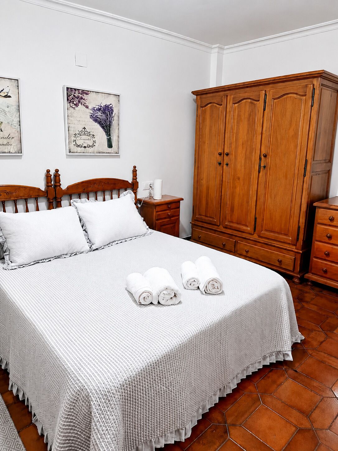 Un lit bien fait, recouvert d’une couvre-lit blanche et agrémenté d’oreillers, une armoire en bois et une table de chevet avec une lampe et une tasse.