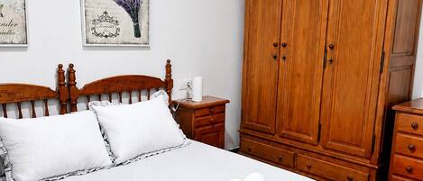 Ein ordentlich bezogenes Bett mit weißer Bettdecke und Kissen, ein Holzschrank und ein Nachttisch mit Lampe und Tasse.