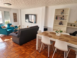 A modern living room with a sofa, a dining table with chairs, a TV mounted on the wall, and a bookshelf. - 🏖️ Spacious 4-bedroom apartment just a short walk from the beach. ☀️ (Vinaròs)