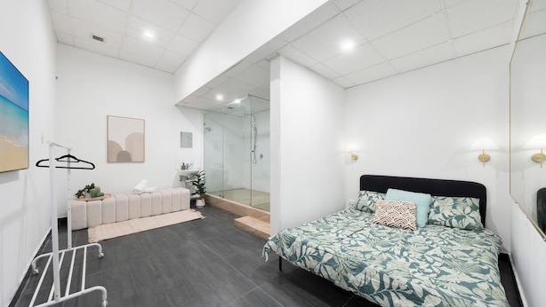 A modern bedroom with a bed, a small seating area, a shower, and a view of the outdoors.