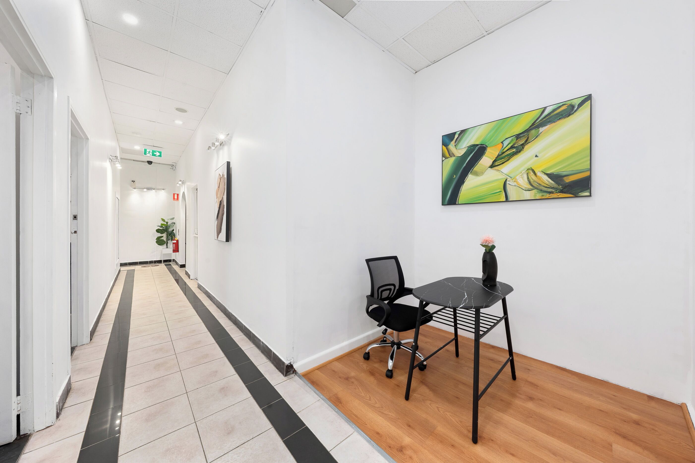 A hallway with a tiled floor, a black office chair, a small table, and a painting on the wall.