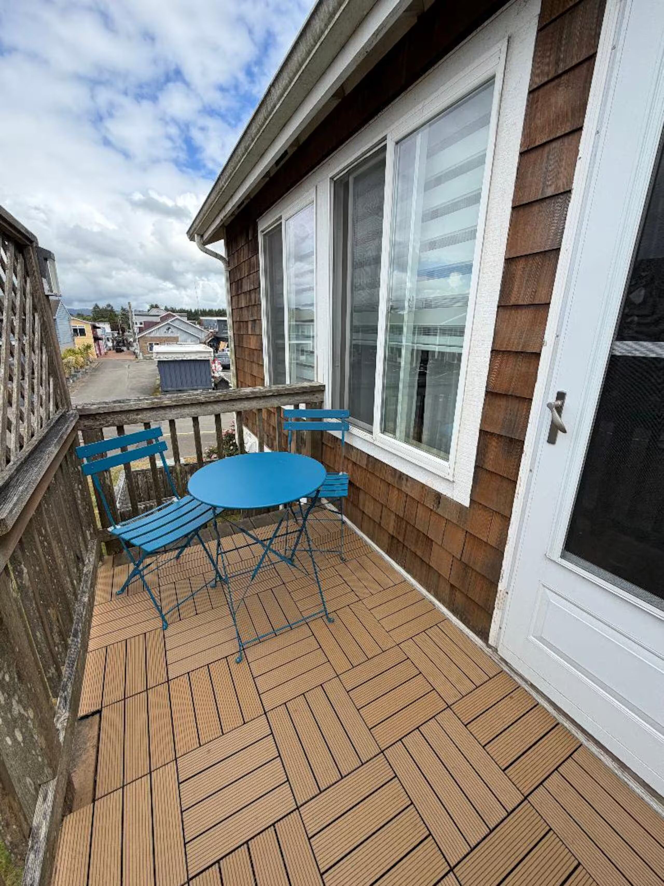 A wooden deck with a blue table and chairs, a white door, and a view of a street.