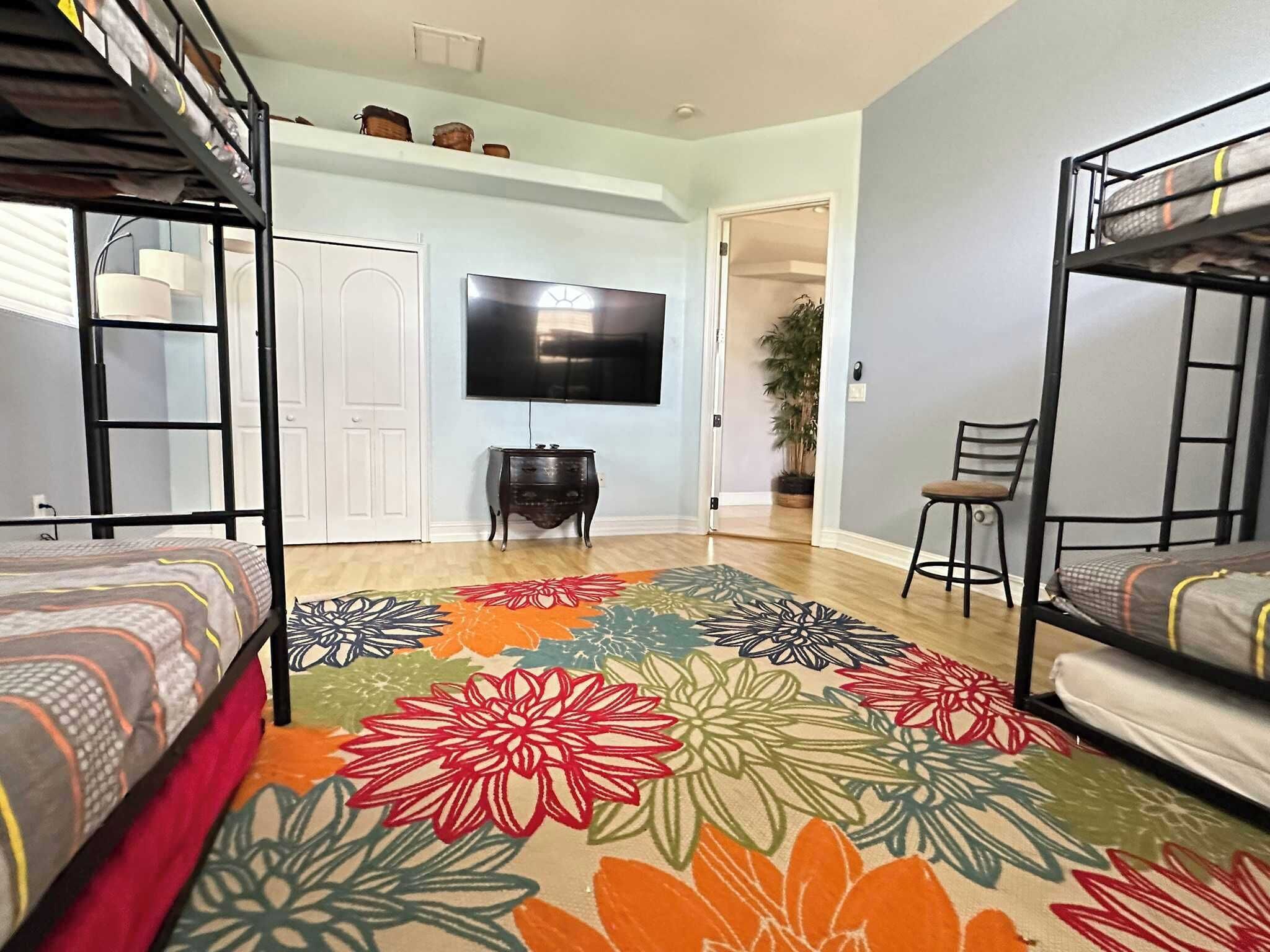 Une chambre de dortoir avec des lits superposés, un téléviseur à écran plat, un bureau et un tapis à motifs floraux aux couleurs vives.