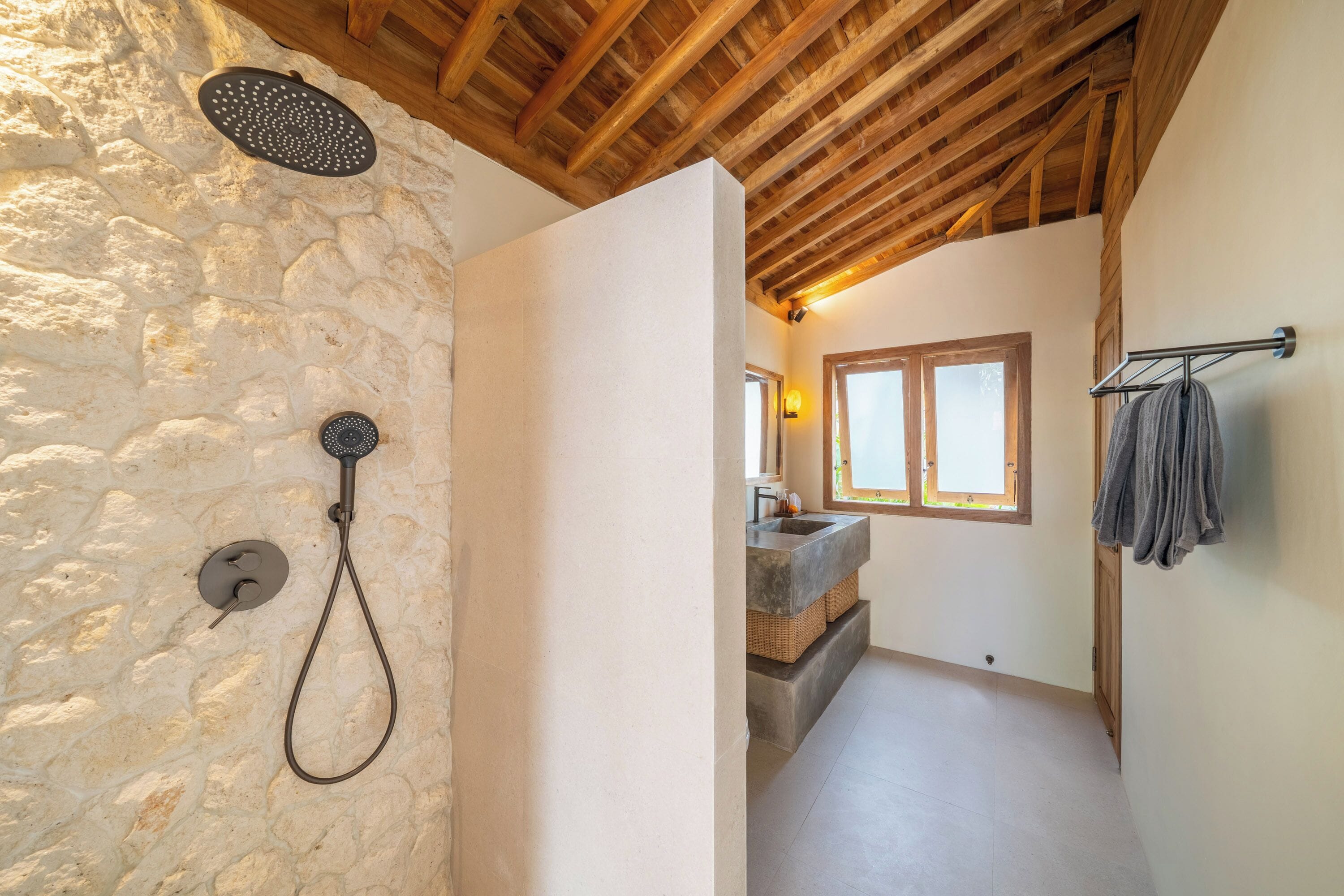 Ein Badezimmer mit Steinsäulen, einer Dusche, einer Holzbalkendecke und Blick in ein Schlafzimmer mit Fenster und Waschbecken.