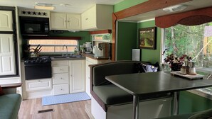 Cozinha compacta de RV com fogão, pia e armários, área de refeições com banco e janela com vista para vegetação.