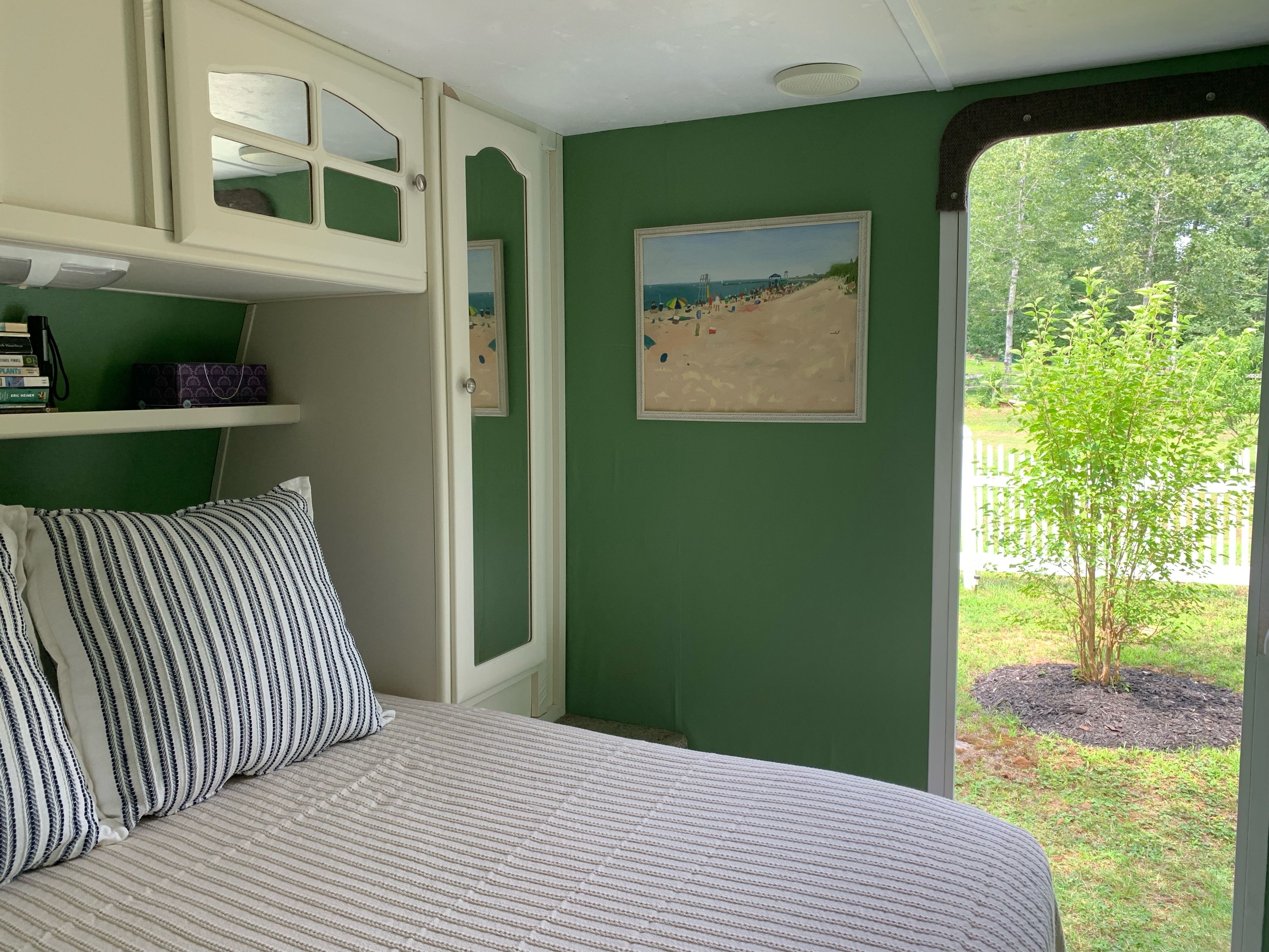 Een slaapkamer met een bed, kussens, een plank met boeken, een ingelijst beeld en uitzicht op een strand door een raam.