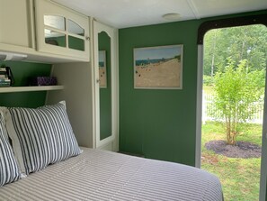 A bedroom with a bed, pillows, a shelf with books, a framed picture, and a view of a beach through a window.