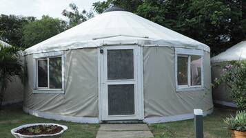 A white, dome-shaped yurt with a door and two windows, surrounded by grass and trees.
