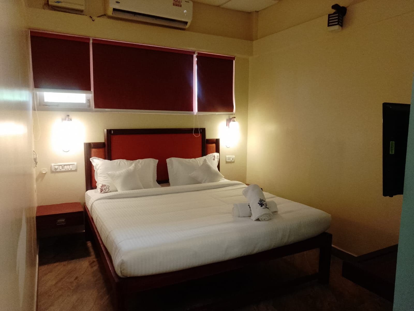 A hotel room with a bed, two bedside tables, a red headboard, and wall-mounted lights.