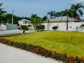 A well-maintained lawn with a garden bed of red flowers, a white building with a satellite dish, and a palm tree. - Spacious 4BR Retreat in El Limón  Mins to Waterfall & AC (El Limón)