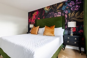 Hotellihuone, jossa on suuri sänky, vihreä pääty ja kaksi yöpöytää, joissa on lamput, sekä kukkakuviotapetti.