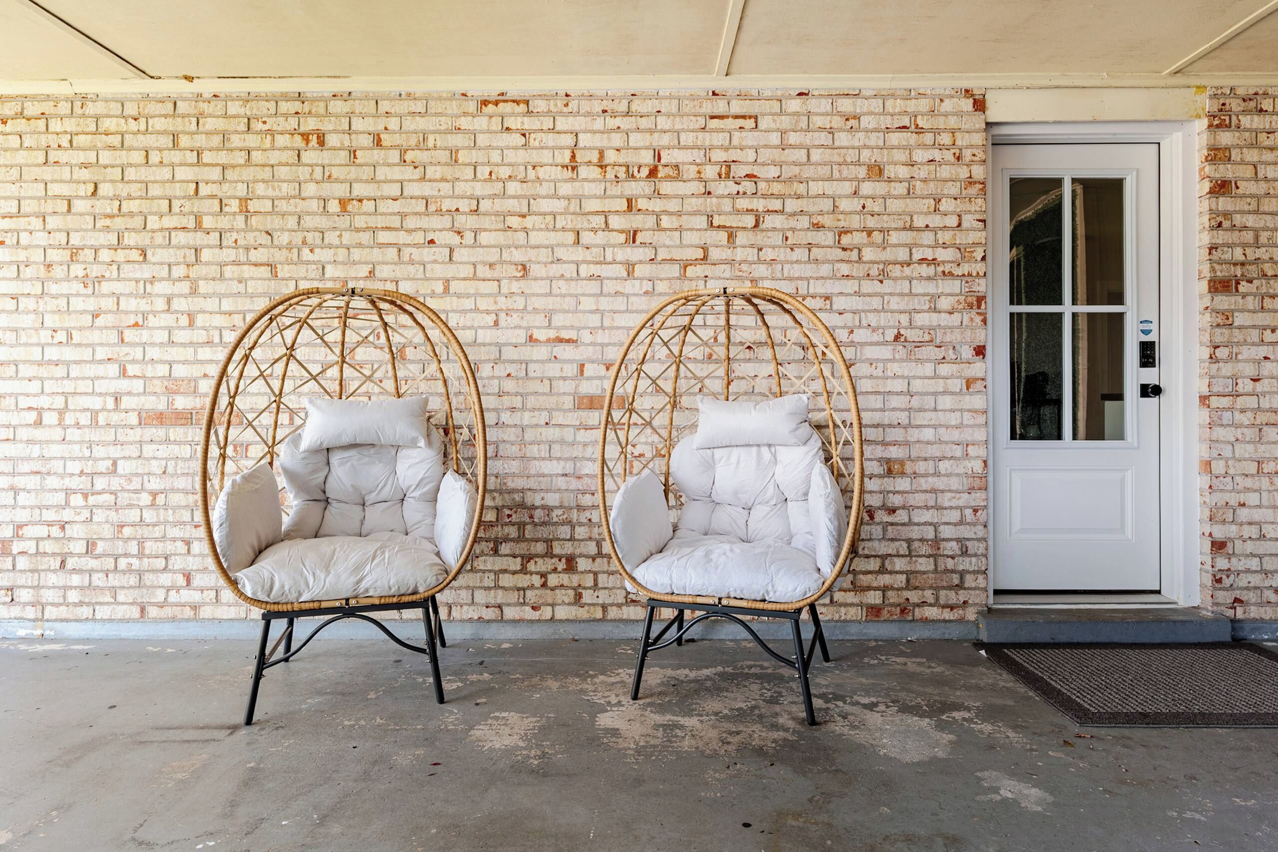 Two wicker chairs with cushions against a brick wall, a white door, and a concrete floor.