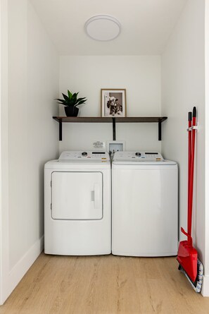 A laundry room with two front-loading washing machines, a shelf with a plant and a framed picture, and a red mop leaning against the wall. - Sleeps 14 • 7 Beds • Airport • Navy Base• Hospitals • Mall • Beaches (Pensacola)