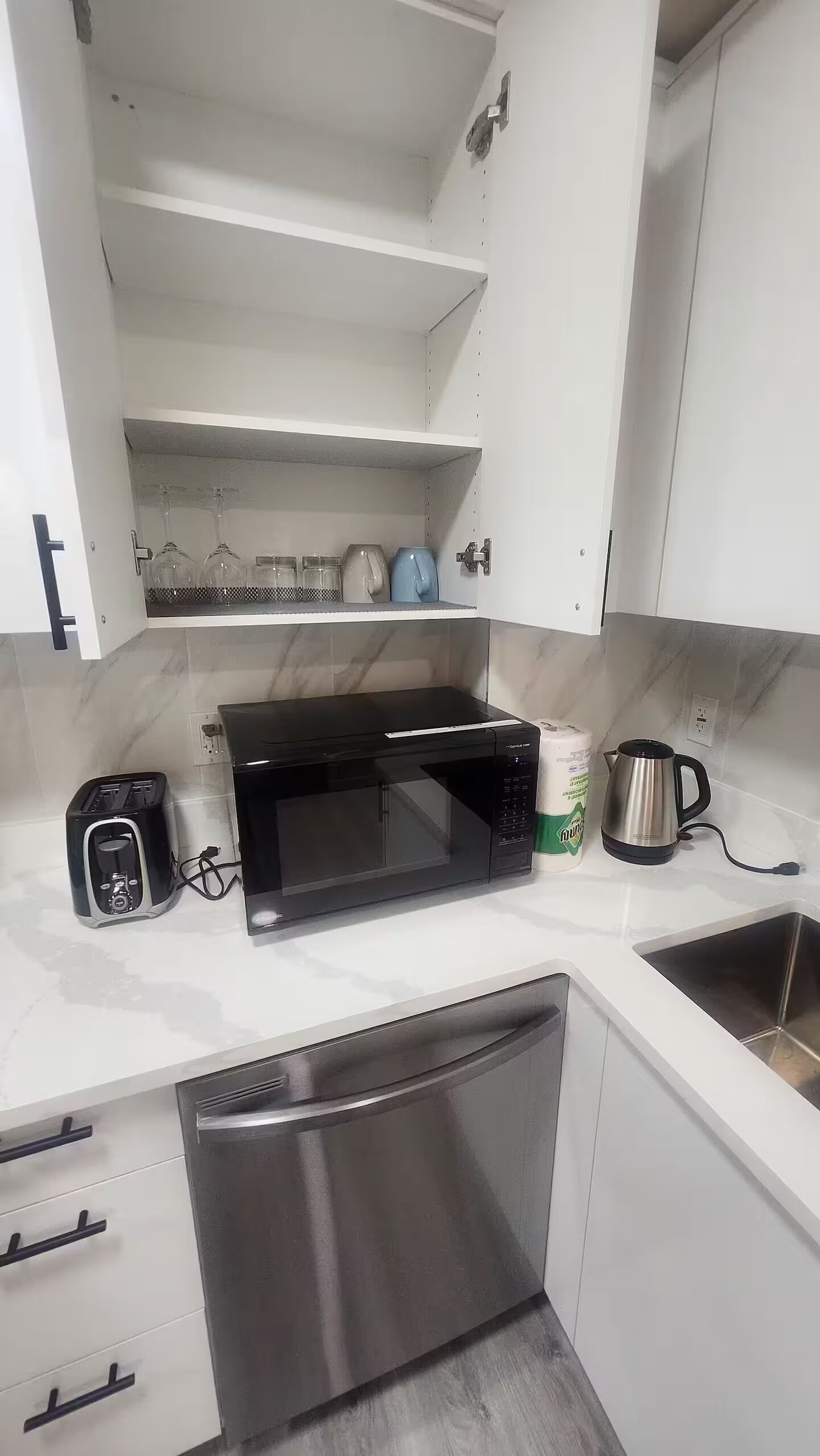 Cocina con armarios blancos, microondas negro, tostadora, hervidor y pileta de acero inoxidable.