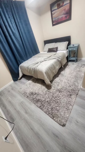 Quarto com cama, mesa de cabeceira, um quadro e cortina azul.