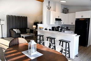 A modern kitchen with white cabinets, a black refrigerator, and a dining area with a wooden table and white chairs. - Enchanting 3BR Soundview | Patio | Firepit | W/D (Camano)