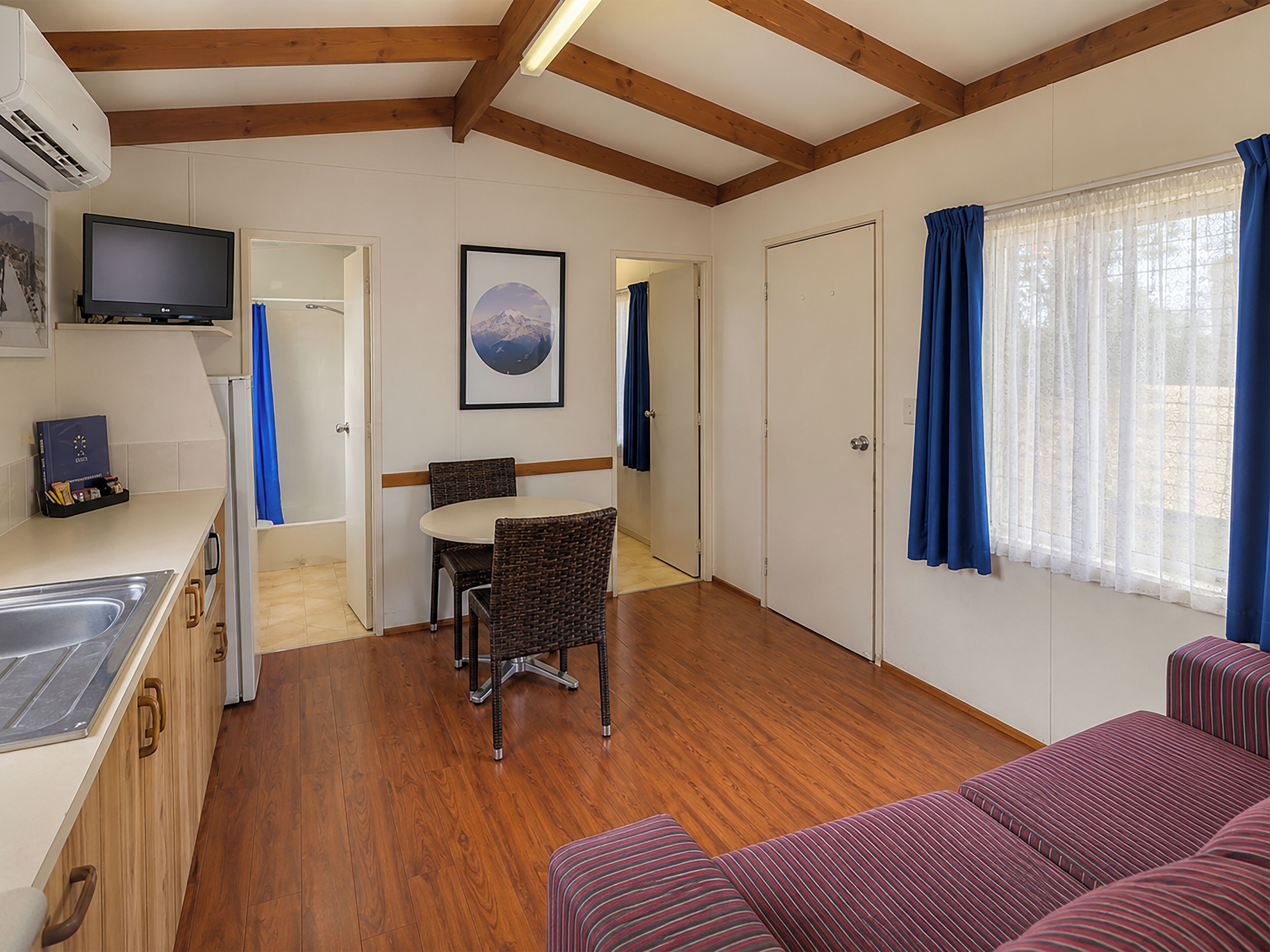 A compact room with a kitchenette, a dining area with a table and chairs, a sofa, and a large window with curtains.