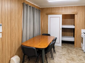 Habitación pequeña con pared de panel de madera, cortina gris, mesa de madera con sillas, puerta blanca y una cama litera.