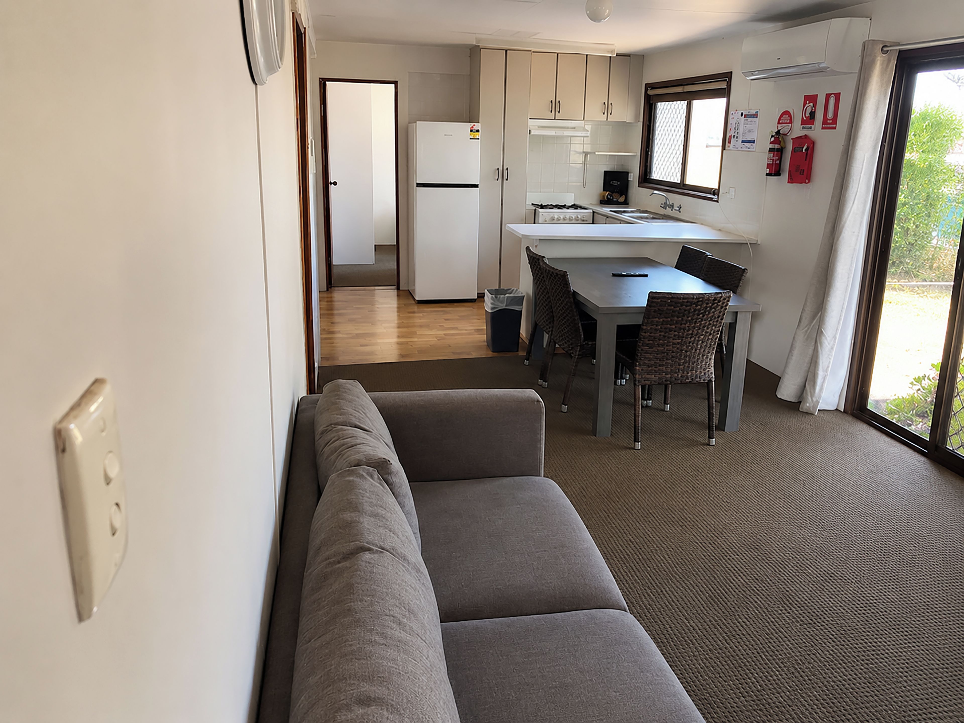 A compact kitchenette with a refrigerator, stove, and sink. There is a dining table with chairs and a bench. The room has a window with curtains and a view of greenery outside.
