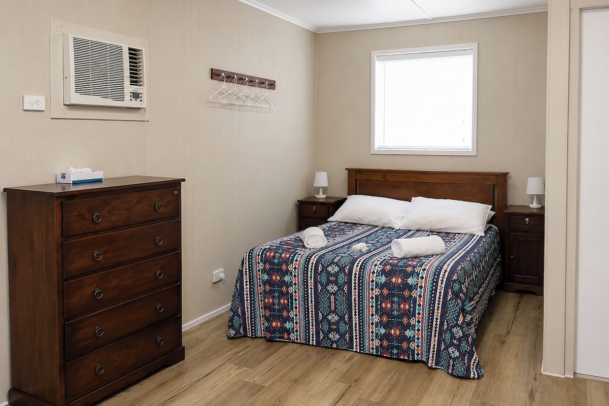 A bedroom with a wooden dresser, a bed with patterned bedding, a window, and a wall-mounted air conditioner.