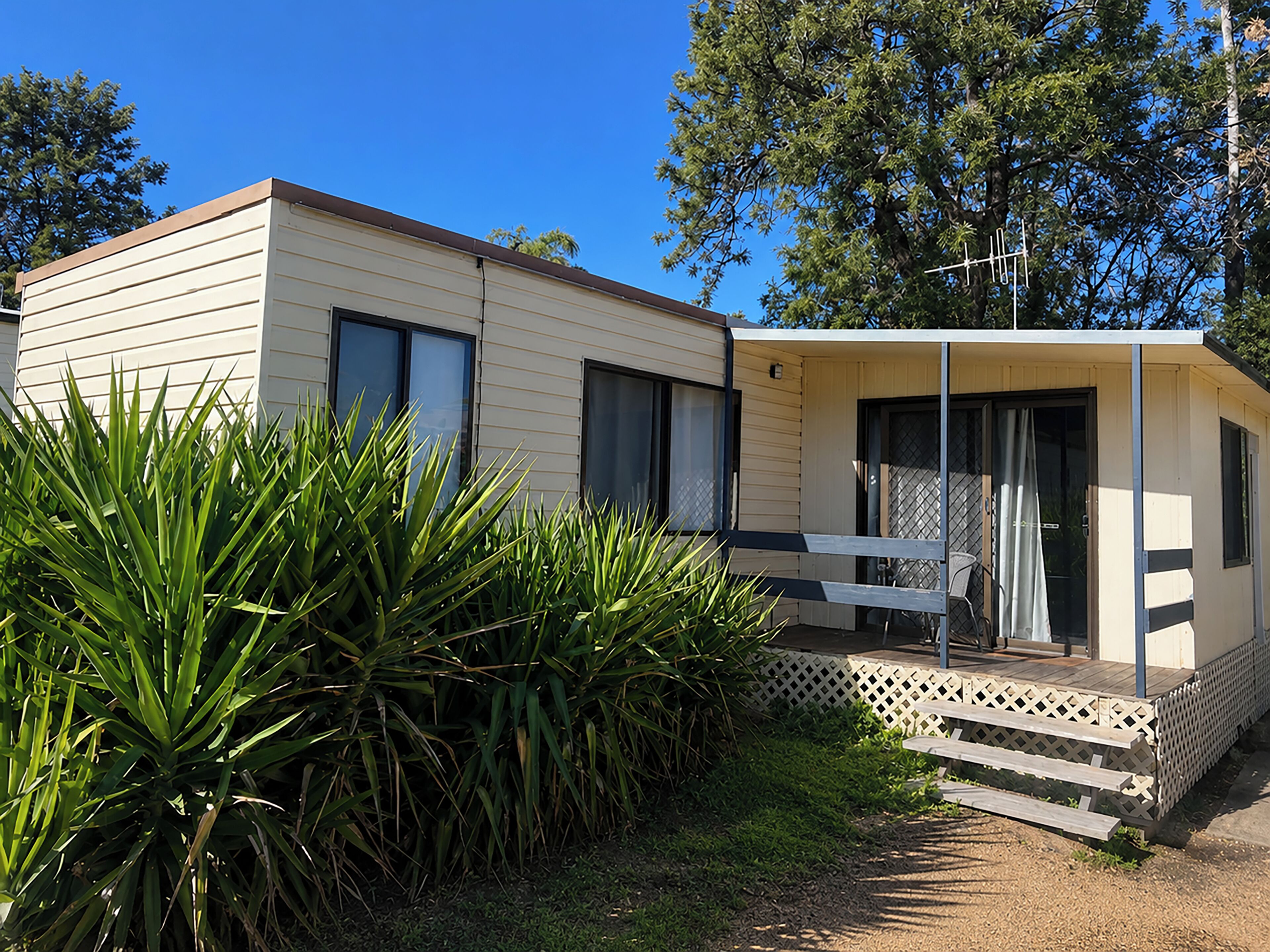 A single-story mobile home with a porch and a small deck, surrounded by greenery.