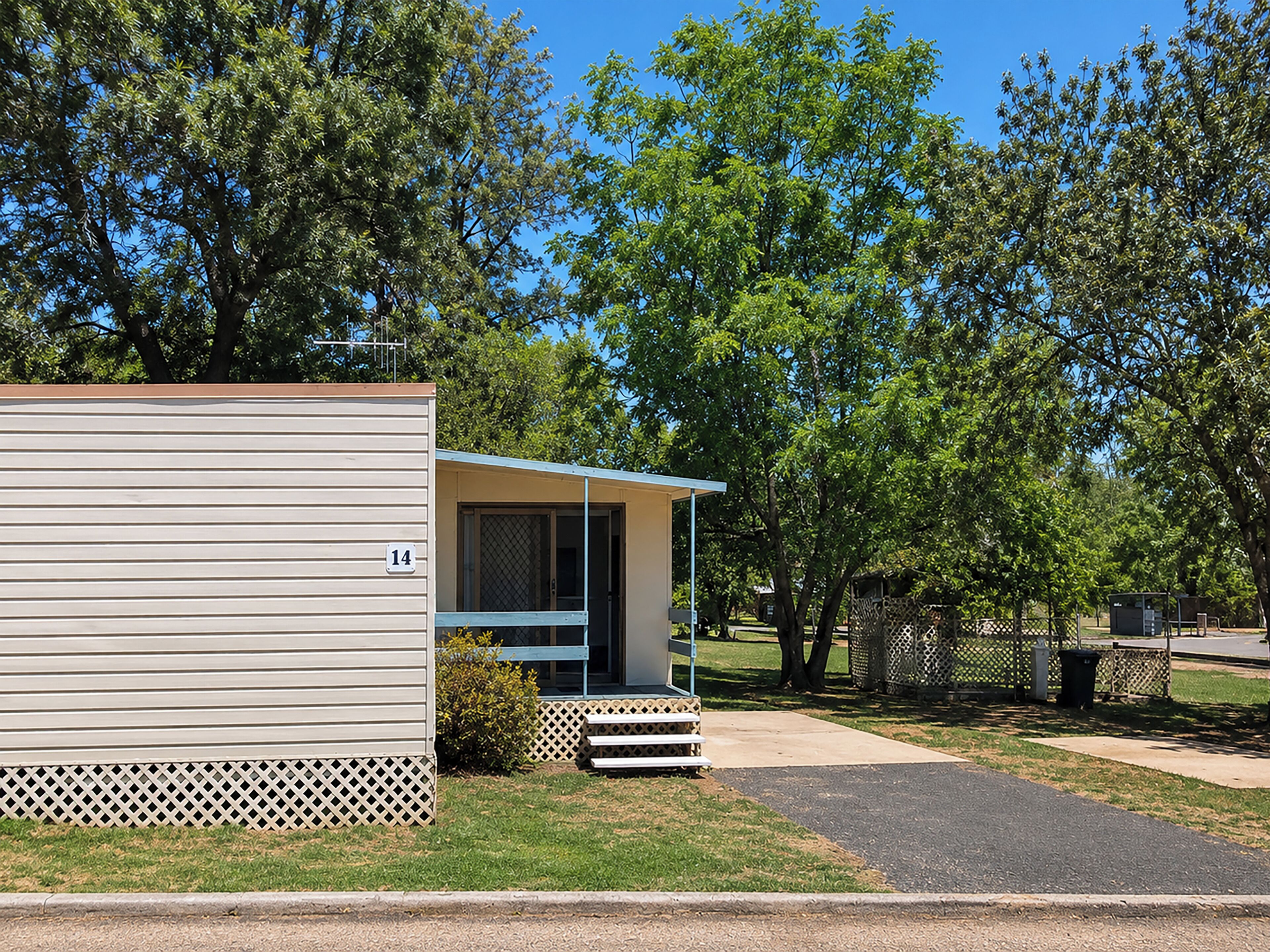A single-story mobile home with a porch, number 14, surrounded by trees and a fenced area.