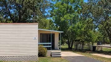 A single-story mobile home with a porch, number 14, surrounded by trees and a fenced area.