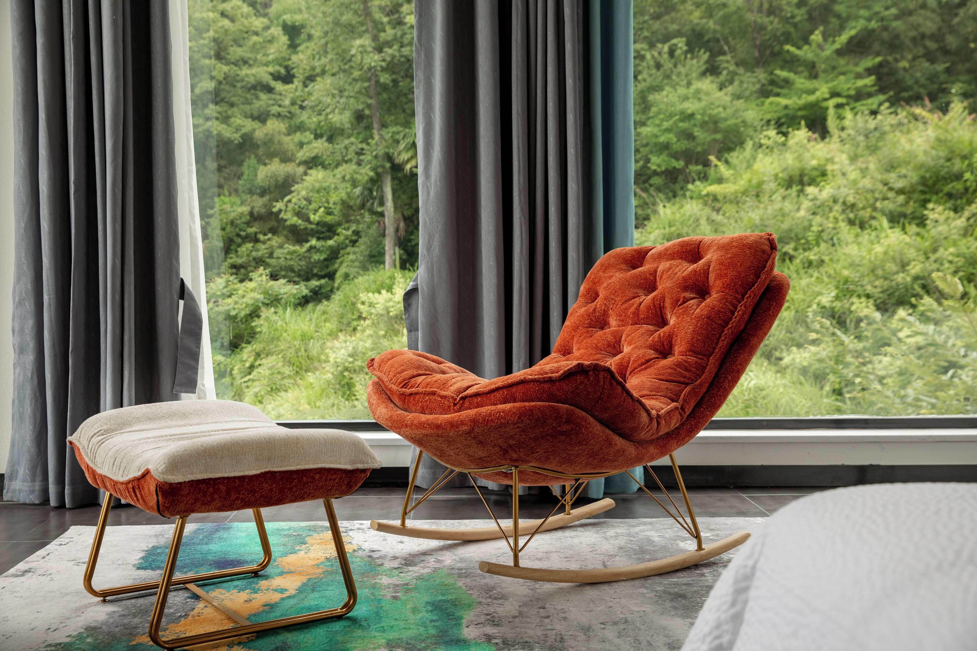 A room with a rocking chair and ottoman by a window with a view of greenery.
