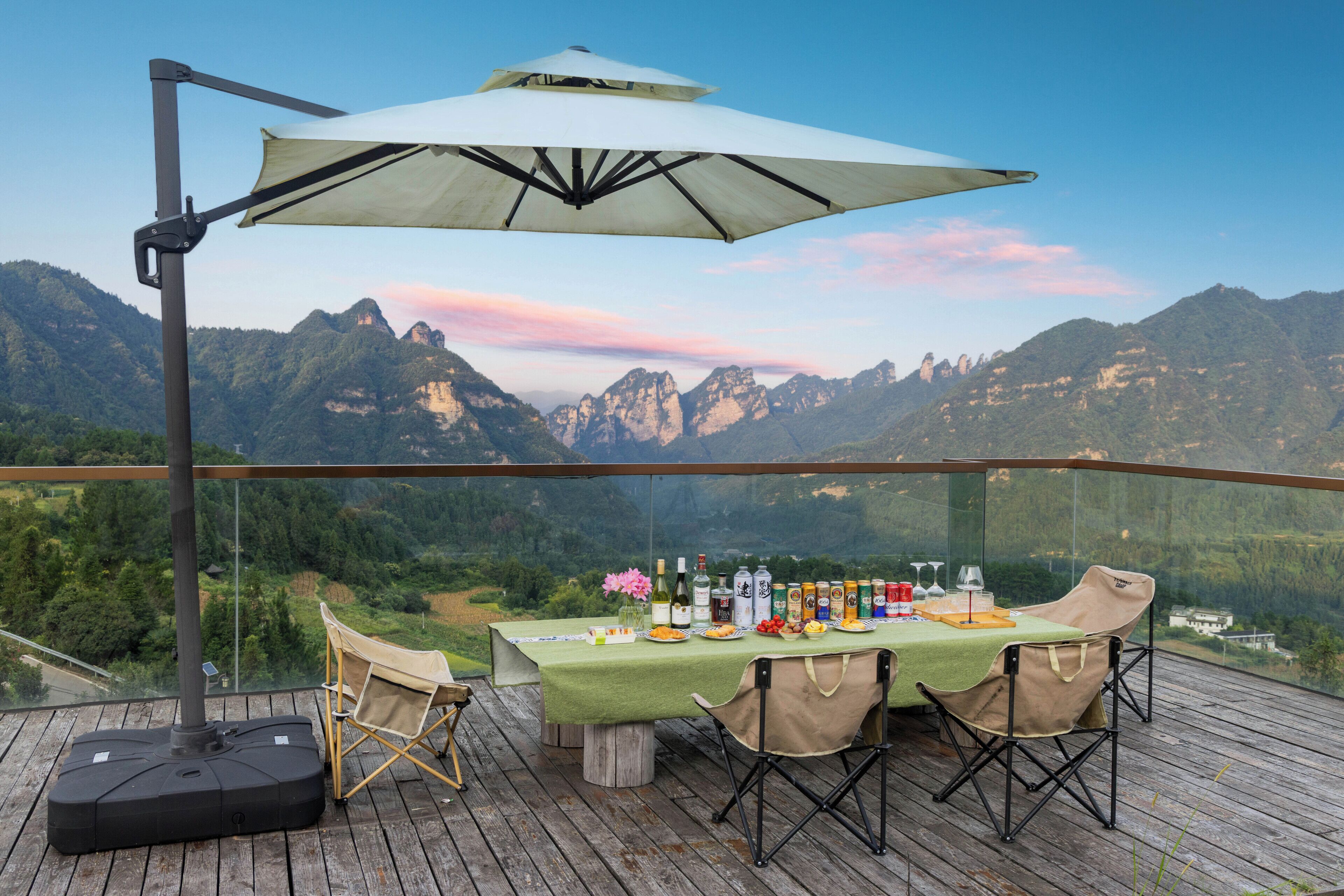 Una mesa de comedor dispuesta en una terraza de madera con vista a las montañas, una lámpara de mesa y diversos alimentos y bebidas.