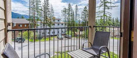 A balcony with two chairs and a table, overlooking a street with parked cars and houses, and a row of tall trees.