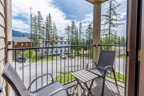 A balcony with two chairs and a table, overlooking a street with parked cars and houses, and a row of tall trees. - Altitude Adjustment | Ski. Shred. Repeat. (Whitefish)