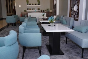 Eine Hotelhalle mit einer langen Reihe blauer Sessel, einem zentralen weißen Tisch und einer Couch mit türkisfarbenen Kissen.