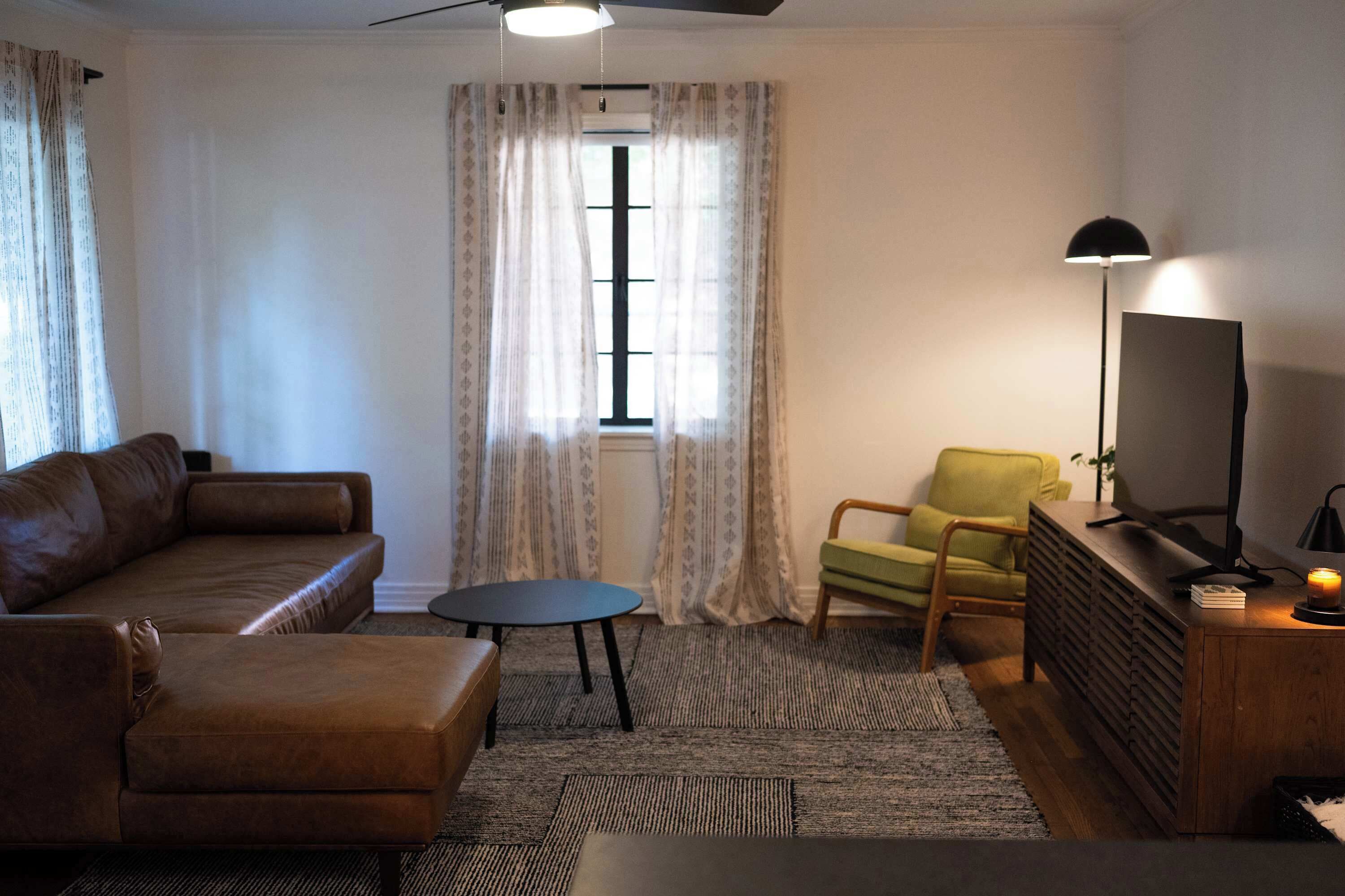 A living room with a brown sofa, a small round table, a green armchair, a TV on a wooden stand, and a ceiling fan.
