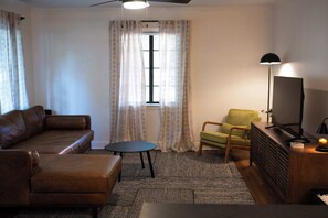A living room with a brown sofa, a small round table, a green armchair, a TV on a wooden stand, and a ceiling fan.