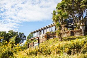 Una casa moderna amb un gran finestral de vidre, envoltada de vegetació i un cel serè.