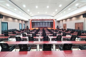A conference room with rows of tables and chairs, a stage with a red backdrop, and a large screen.