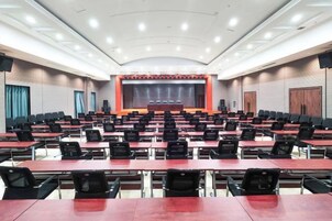 Un salón de conferencias con filas de mesas y sillas, un escenario con un telón rojo y una pantalla grande.