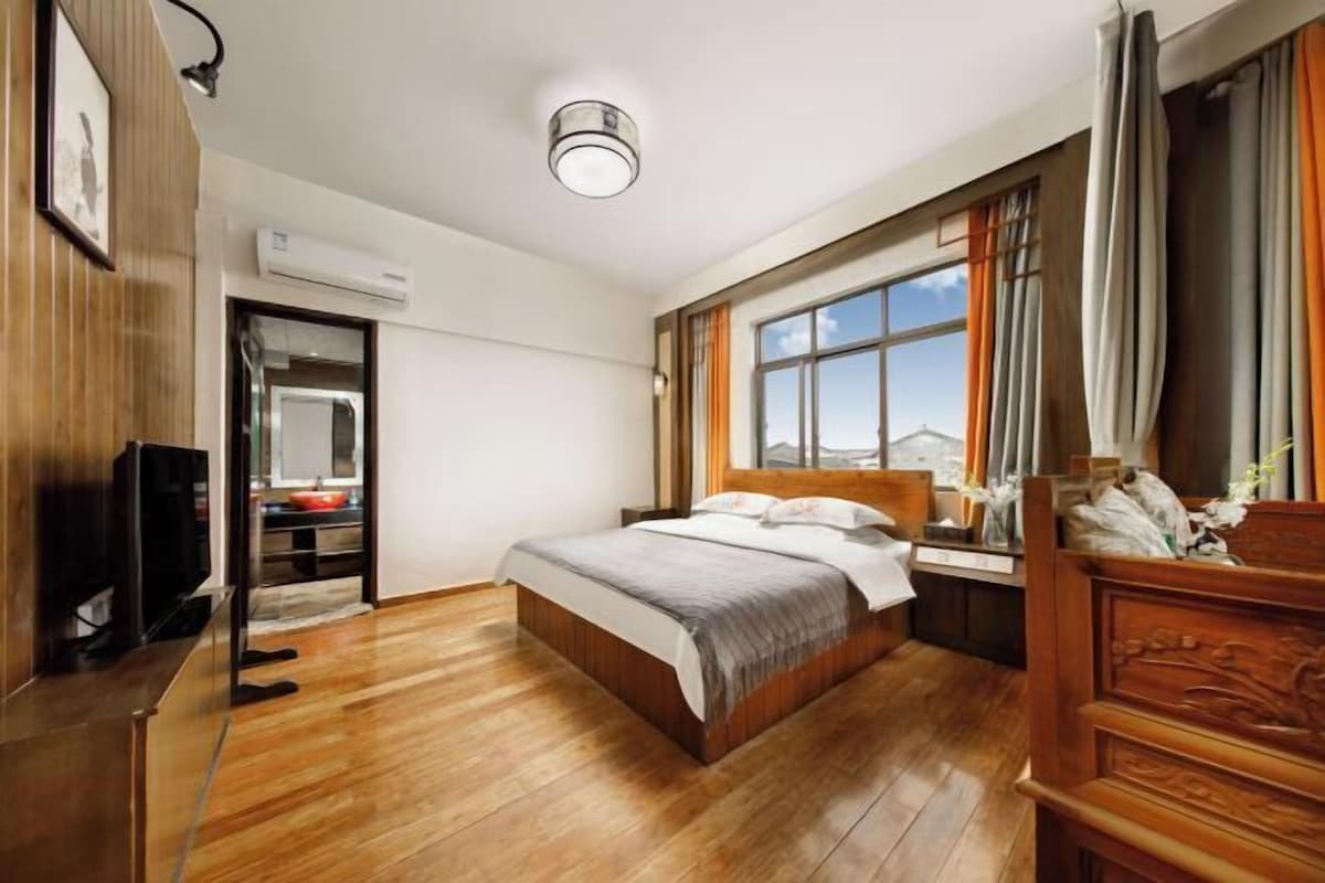 A hotel room with a wooden floor, a bed with a headboard, a TV, a window with curtains, and a desk with a chair.