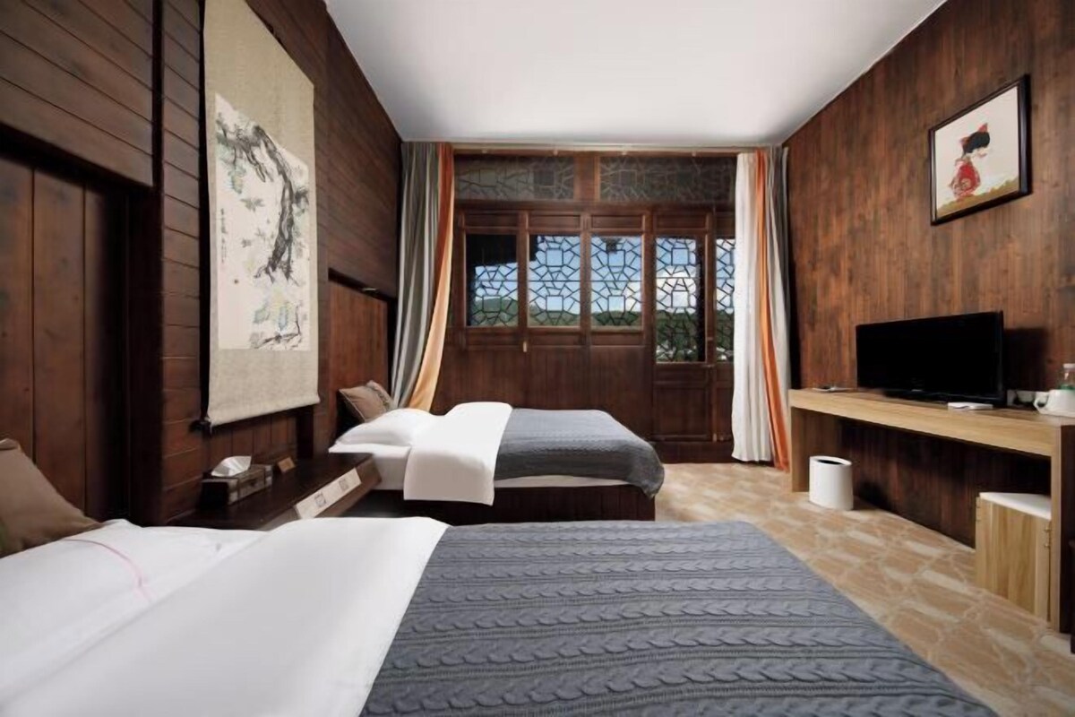 A room with two beds, a television, wooden walls, a window with curtains, and a painting.