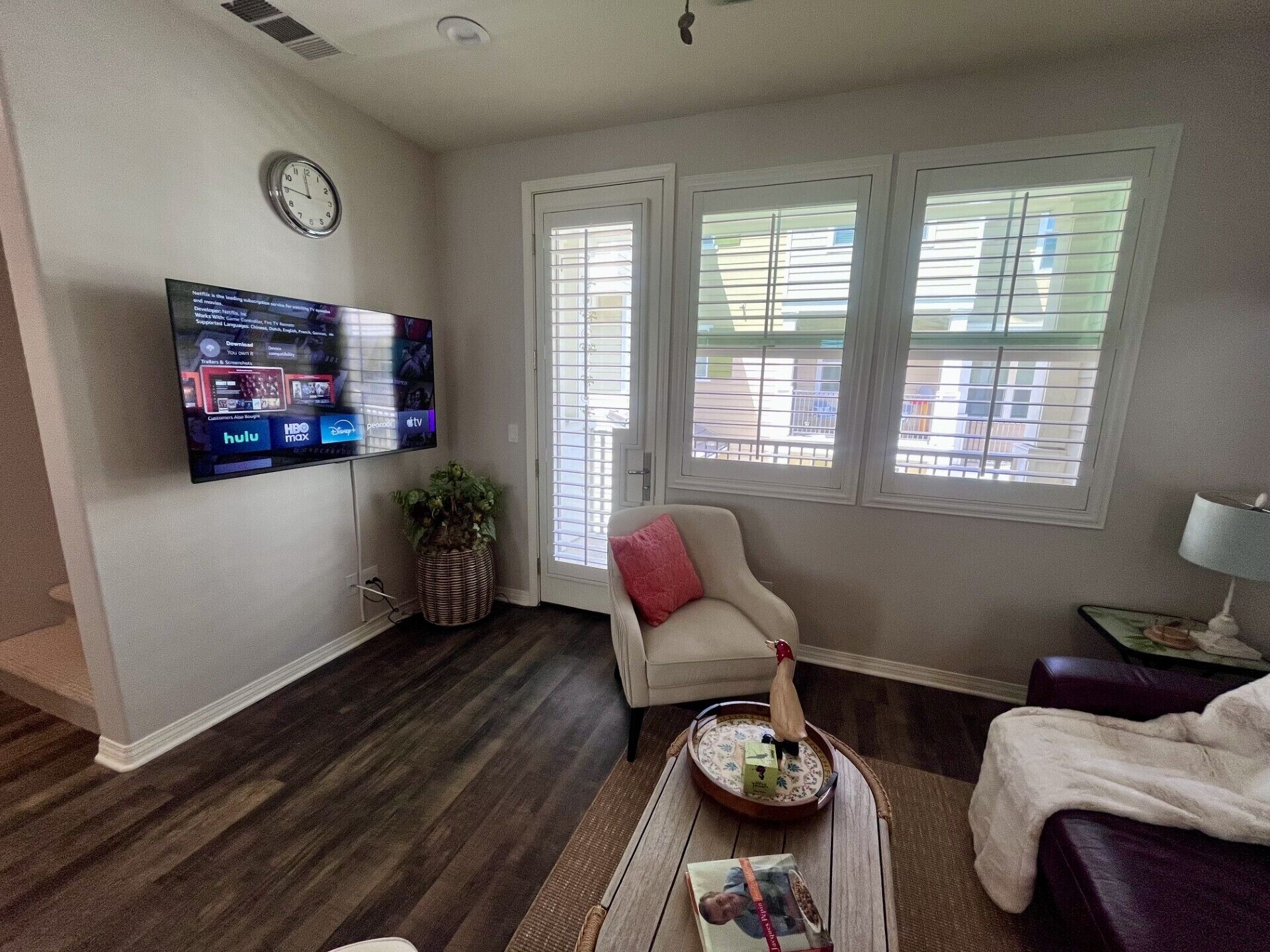 A living room with a flat-screen TV mounted on the wall, a comfortable armchair, a small table with a tray, and a window with blinds.