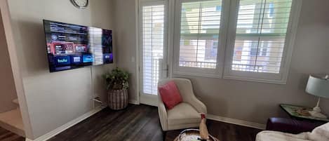 A living room with a flat-screen TV mounted on the wall, a comfortable armchair, a small table with a tray, and a window with blinds.