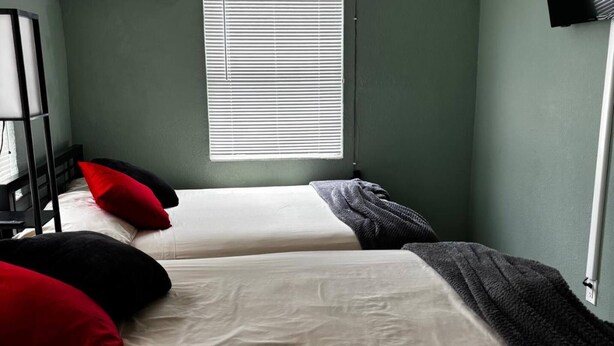 A room with two beds, each with white bedding and red and black pillows. There is a window with blinds, a wall-mounted television, and a power outlet on the wall. - Downtown Sarasota 2BR Apartment 2BA Pet Friendly Beaches Sleeps 5 (Sarasota)