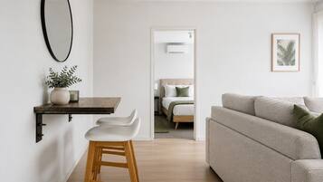 Un salon moderne avec un canapé, un tabouret en bois, une petite table sur laquelle est posée une plante, et un tableau encadré accroché au mur.