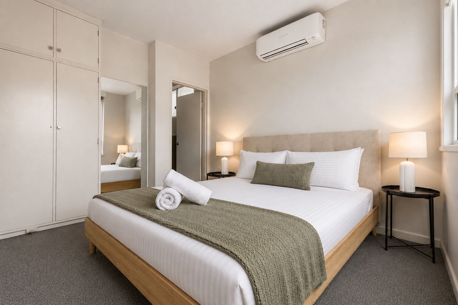 A hotel room with a bed, bedside lamps, an air conditioning unit, and a wardrobe.
