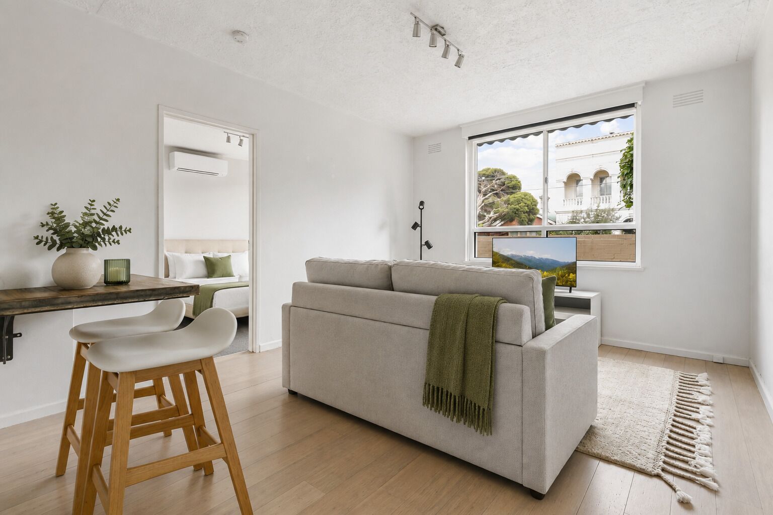 A modern living room with a sofa, a dining area with a table and chairs, and a large window with a view of buildings and trees.