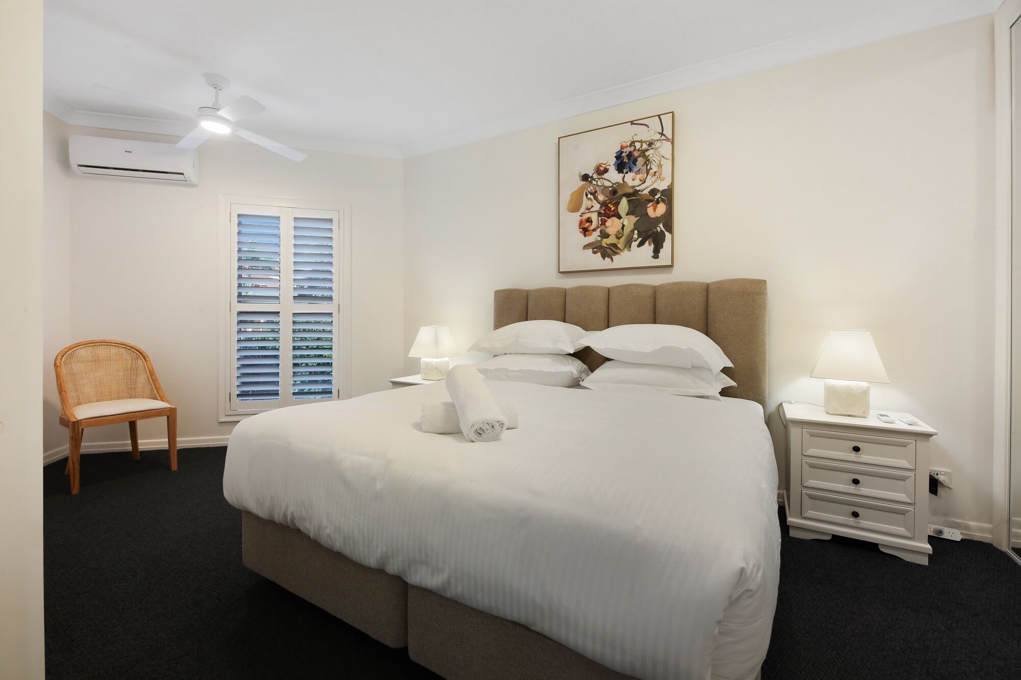 Une chambre d’hôtel comprenant un lit, deux tables de chevet, une chaise, une fenêtre avec stores, un appareil de climatisation et un ventilateur de plafond.