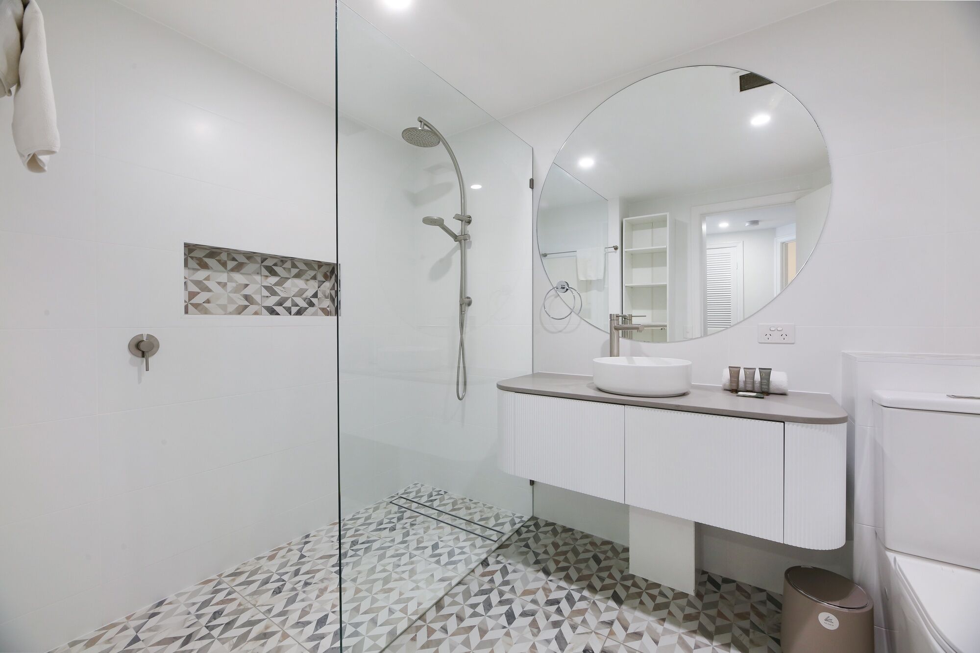 Un baño moderno con ducha de mampara de vidrio, un espejo redondo y un lavabo blanco.