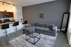 A modern living room with a grey sofa, two black bar stools, a glass coffee table, and a kitchen area with a microwave and stove.